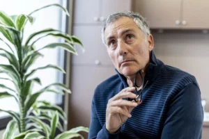 Homme d'âge mûr réfléchi tenant ses lunettes devant une plante verte.