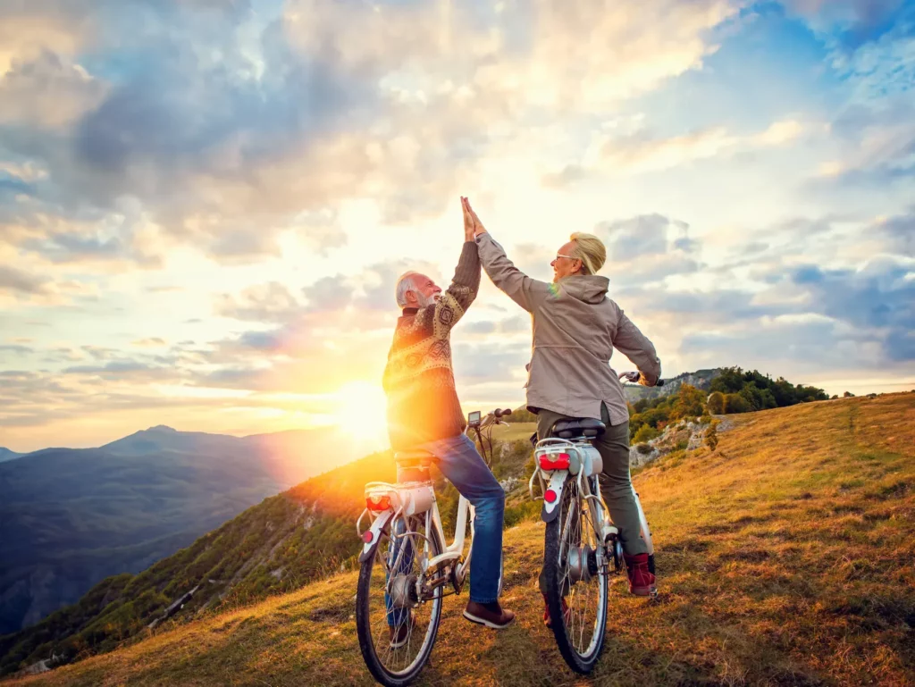 Couple de seniors à vélo se félicitant d'un high-five en haut d'une colline au coucher du soleil.