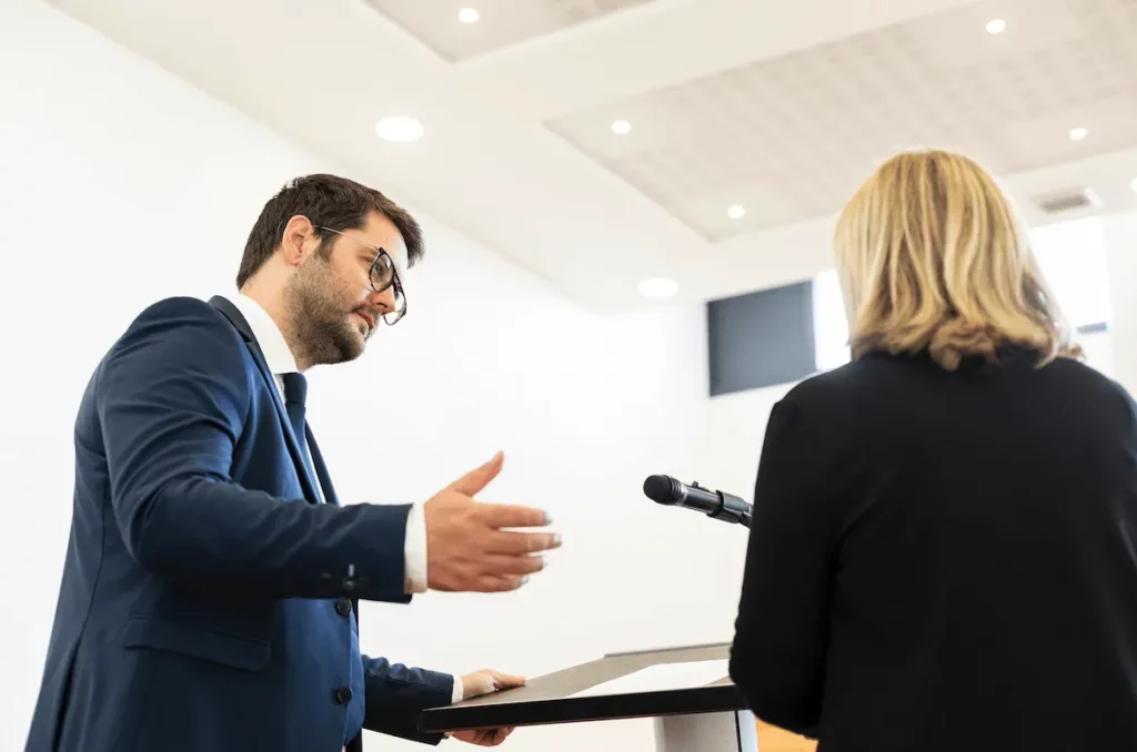 Orateur s'exprimant avec un geste de la main lors d'une cérémonie funéraire en intérieur.